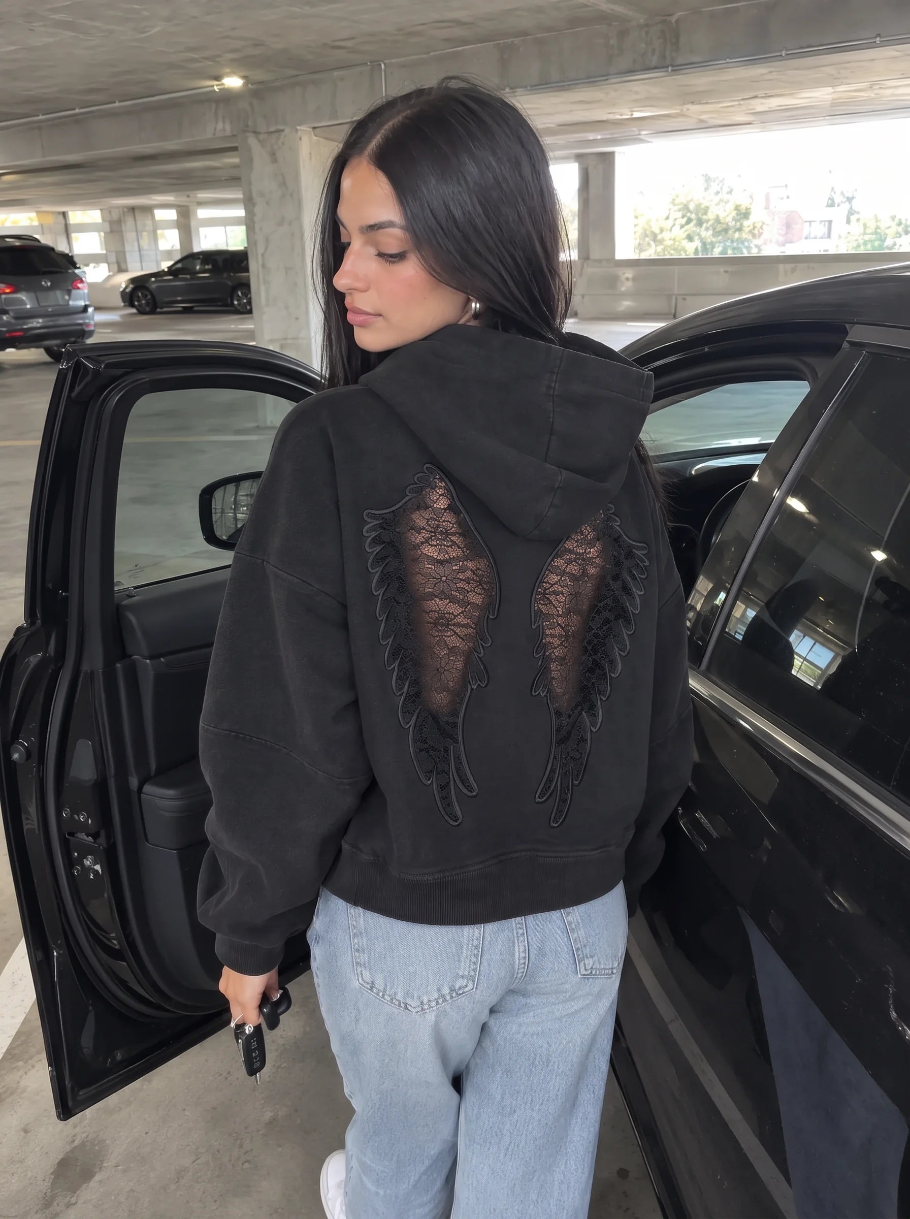 Black Laced Wing Hoodie with a soft downward glance in a parking garage.